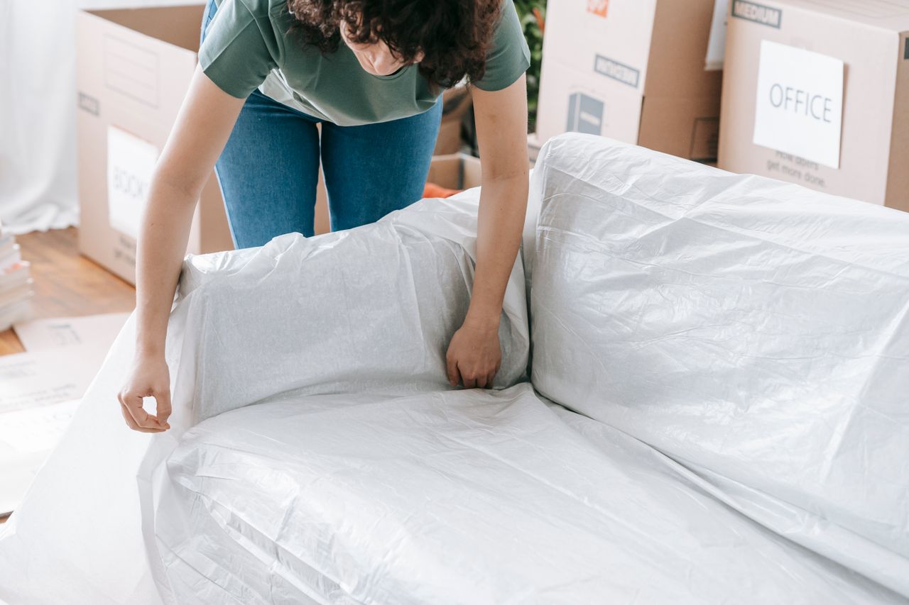 services-03 A woman carefully wraps a sofa in protective material while packing boxes in a bright room.